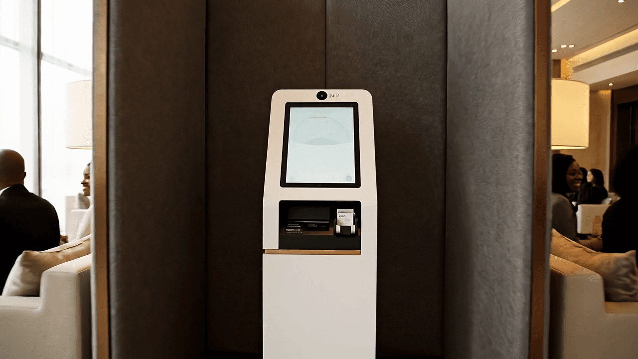 Belebte Kamerafahrt: durch den Hoteleingang in eine lebendige Lobby mit Lounge-Bereich, in dem sich Gäste auf Sofas unterhalten; vorbei an einem akustisch abgeschirmten Self-Service-Bereich mit dem ECG AI Terminal, dessen Display einen freundlichen Avatar mit sichtbarem Gesicht und den Gruß „Herzlich Willkommen" zeigt; abschließend ein Schwenk zum besetzten Empfang, an dem eine Mitarbeiterin Gäste eincheckt, während sich feine champagner-bronzene Verbindungslinien zeichnen.